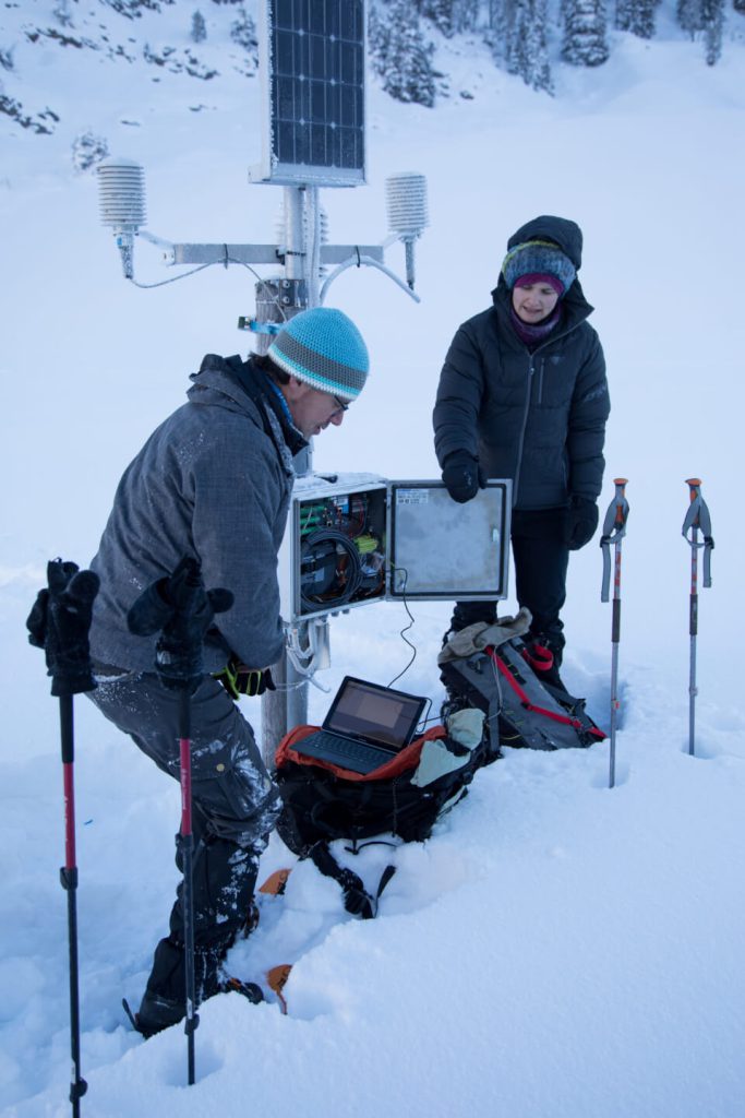 Zwei Personen warten im Schnee eine Wetterstation mit Solarpanel, Laptop und Schneeschuhen