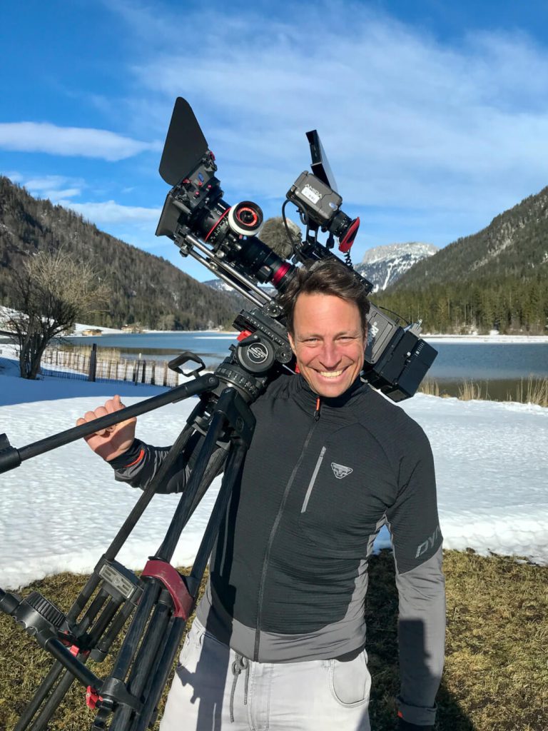 Kameramann trägt Profikamera mit Stativ, verschneite Landschaft, Videoproduktion Outdoor