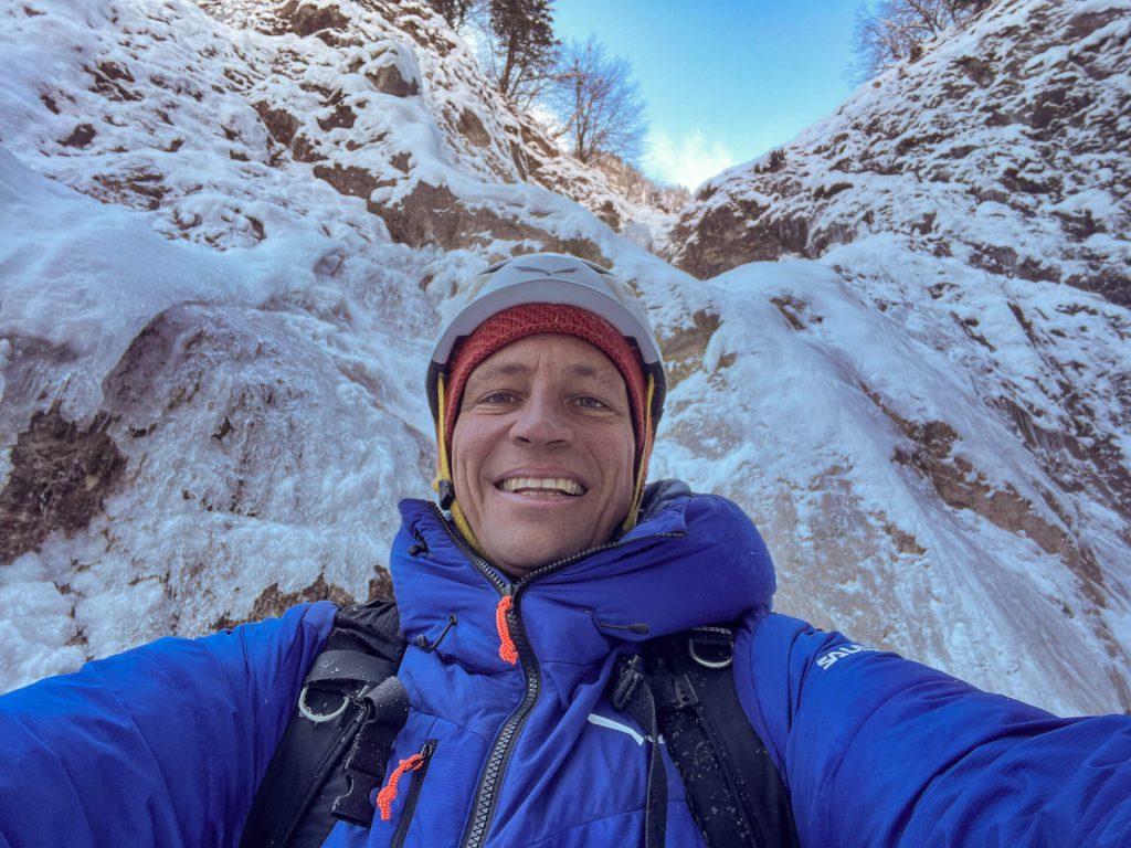 Mann mit Helm und blauer Jacke macht Selfie beim Winterwandern in verschneiter Berglandschaft