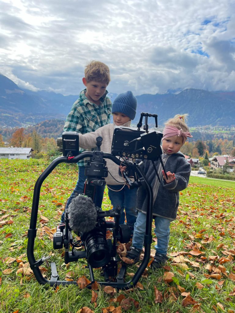Drei Kinder bedienen professionelle Filmkamera auf Wiese vor Alpenpanorama, Videoproduktion