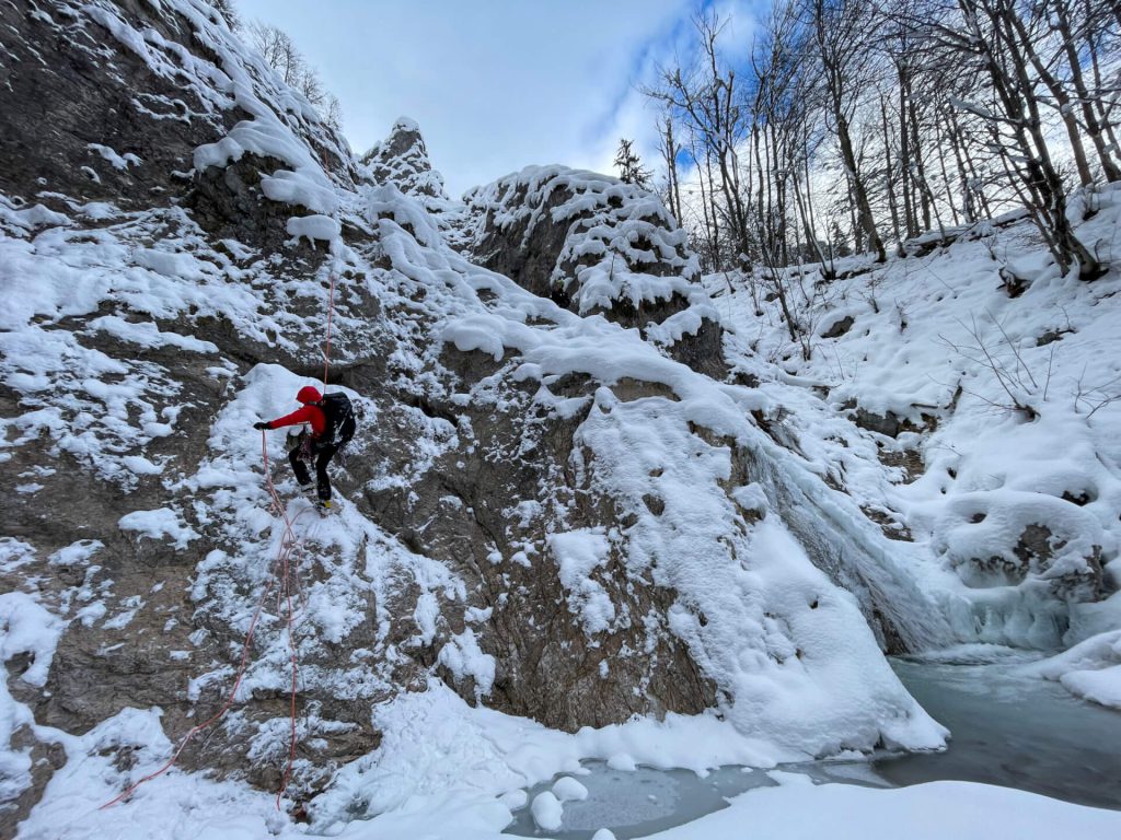 Bergsteiger mit roter Jacke klettert im Winter an schneebedecktem Felsen, Drohnenaufnahme