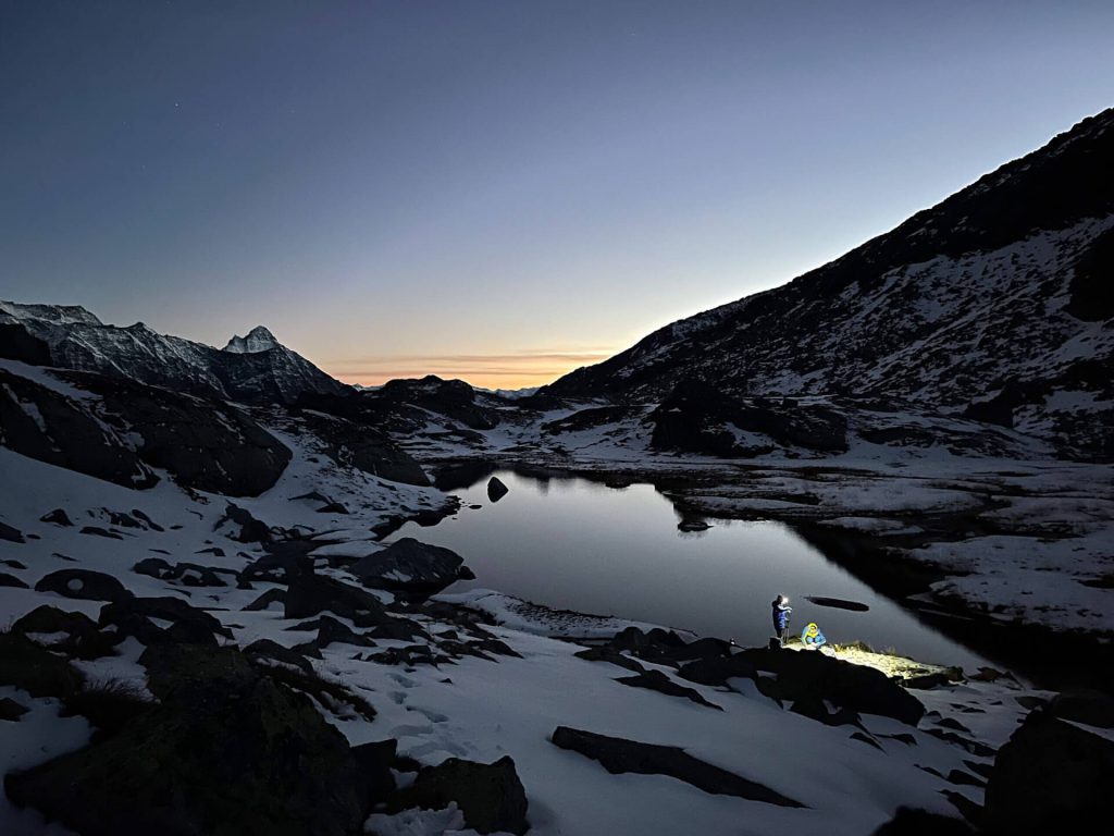 Schneebedeckte Berge bei Sonnenuntergang, Drohnenaufnahme mit Mensch und Beleuchtung am Seeufer
