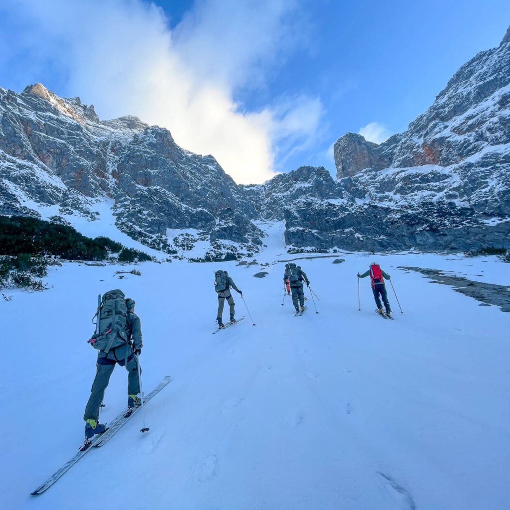 Vier Skitourengeher steigen bei Sonnenschein durch verschneite Berglandschaft Richtung Gipfel